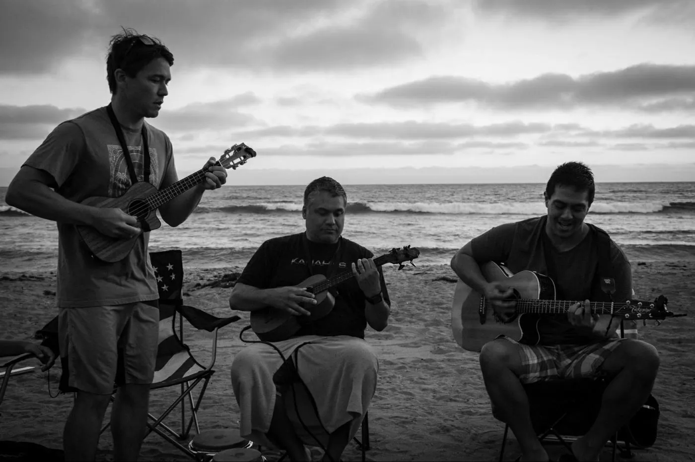 Three musicians playing ukulele and guitar on a beach at dusk